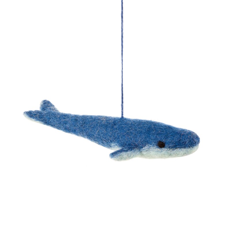 Op de afbeelding: Een blauwe vilten walvis-versiering die aan een touwtje hangt. De walvis heeft een lichtblauwe buik en zwarte ogen.