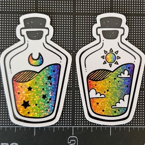 May include: Two iridescent glitter filled glass bottles with cork stoppers. One bottle has a crescent moon and stars inside, the other has a sun and clouds inside.