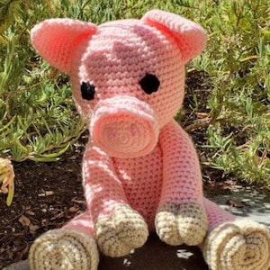 May include: A pink crocheted pig toy with black eyes and beige hooves. The pig is sitting on a gray surface.