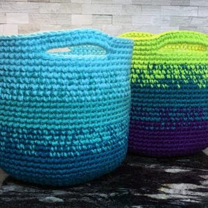 May include: Two crocheted baskets with ombre color patterns. The basket on the left is blue and green, and the basket on the right is green, blue, and purple.
