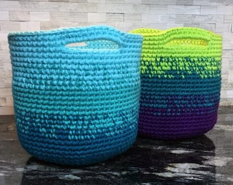 May include: Two crocheted baskets with ombre color patterns. The basket on the left is blue and green, and the basket on the right is green, blue, and purple.