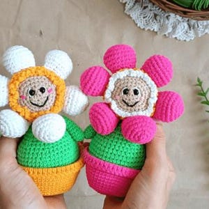 May include: Two crocheted flower pot toys. One is yellow and white with a green pot. The other is pink with a green pot.