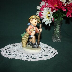 May include: A figurine of a young boy wearing a brown hat and carrying a bundle of sticks. He is wearing a blue shirt and brown pants. The figurine is standing on a white doily.