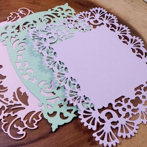 Puede incluir: Tres recortes de papel decorativos en colores pastel: rosa claro, verde menta y lavanda. Cada recorte tiene un borde detallado y ornamentado con dise&ntilde;os florales y de volutas. Los recortes est&aacute;n superpuestos sobre una superficie de madera.