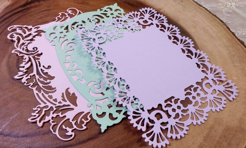 Puede incluir: Tres recortes de papel decorativos en colores pastel: rosa claro, verde menta y lavanda. Cada recorte tiene un borde detallado y ornamentado con dise&ntilde;os florales y de volutas. Los recortes est&aacute;n superpuestos sobre una superficie de madera.
