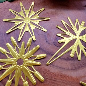 Op de afbeelding: Close-up van verschillende goudkleurige metalen versieringen in de vorm van een sterrenuitbarsting. Elke sterrenuitbarsting heeft een uniek ontwerp met puntige stralen en ingewikkelde uitgesneden patronen. De objecten zijn gerangschikt op een donker houten oppervlak.
