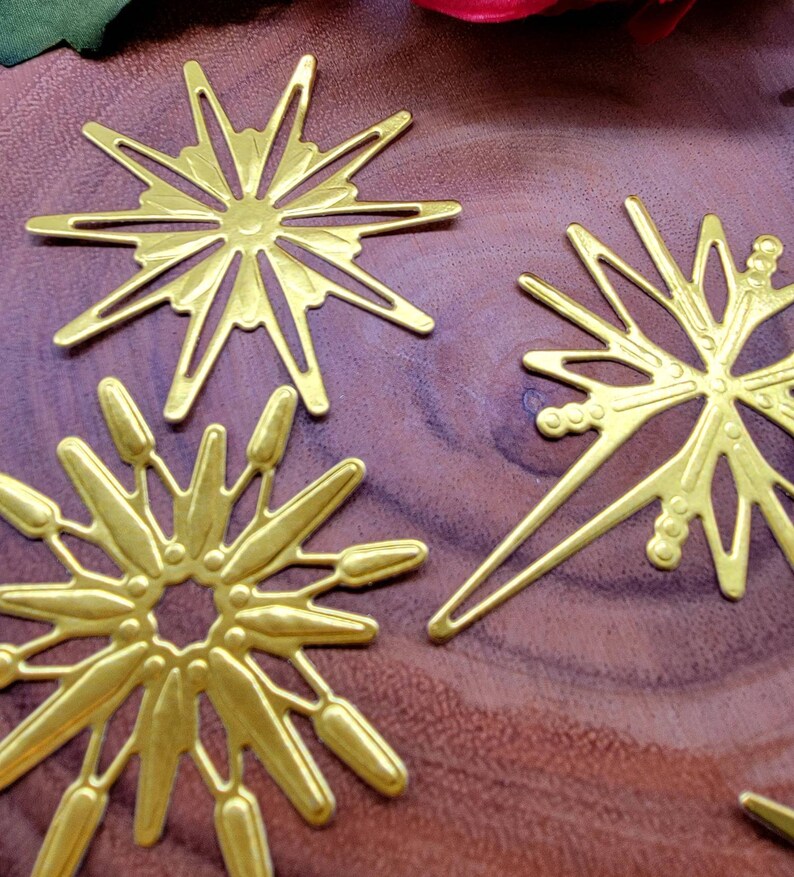 Op de afbeelding: Close-up van verschillende goudkleurige metalen versieringen in de vorm van een sterrenuitbarsting. Elke sterrenuitbarsting heeft een uniek ontwerp met puntige stralen en ingewikkelde uitgesneden patronen. De objecten zijn gerangschikt op een donker houten oppervlak.