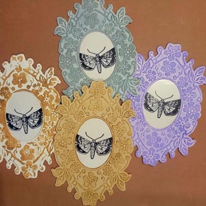 May include: Four decorative oval frames in shades of gold, gray, and purple, each with a detailed floral border. Inside each frame is a black and white illustration of a moth. The frames are arranged on a brown background.