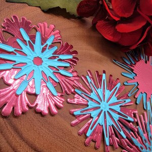 May include: Close-up of several decorative snowflakes in shades of pink and blue. The snowflakes have a layered design, with a pink base and a bright blue center. The background includes red flowers and a green leaf.
