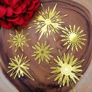 Op de afbeelding: Een verzameling van zes goudkleurige sterrenburst ornamenten met ingewikkelde uitgesneden ontwerpen. De ornamenten vari&euml;ren in grootte en vorm, met puntige en afgeronde randen. Ze zijn gerangschikt op een houten oppervlak, met een tros rode bloemen linksboven.