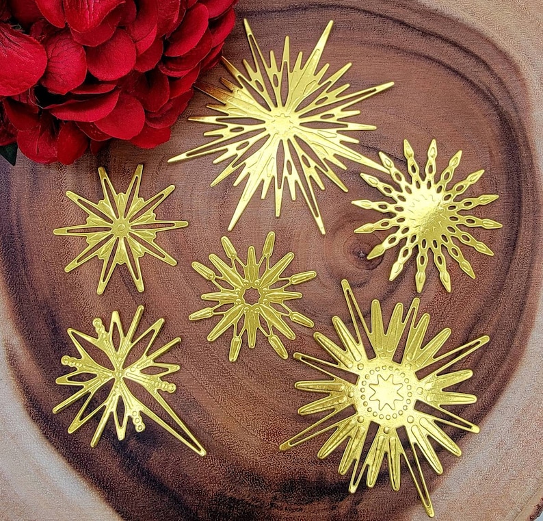 Op de afbeelding: Een verzameling van zes goudkleurige sterrenburst ornamenten met ingewikkelde uitgesneden ontwerpen. De ornamenten vari&euml;ren in grootte en vorm, met puntige en afgeronde randen. Ze zijn gerangschikt op een houten oppervlak, met een tros rode bloemen linksboven.