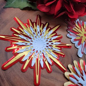 Op de afbeelding: Close-up van decoratieve steruitbarstingsornamenten in rood, goud en wit. De steruitbarstingen hebben een gelaagd ontwerp met een metallic afwerking. De achtergrond bevat rode bloemen en een houten oppervlak.