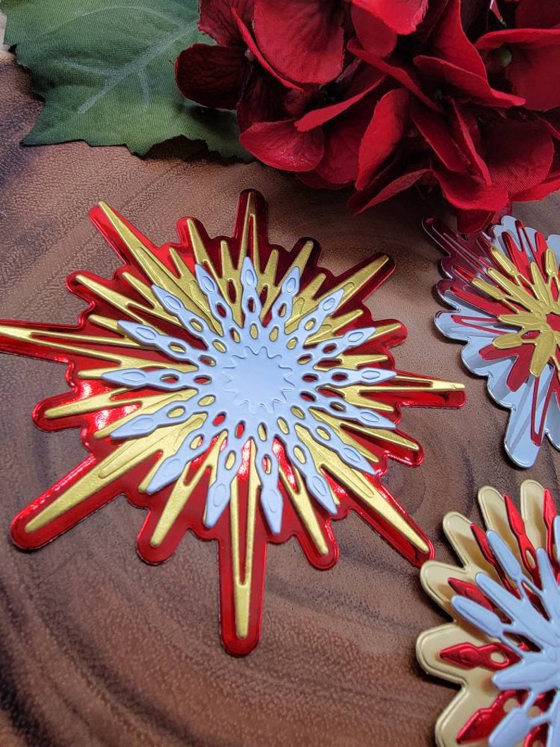 Op de afbeelding: Close-up van decoratieve steruitbarstingsornamenten in rood, goud en wit. De steruitbarstingen hebben een gelaagd ontwerp met een metallic afwerking. De achtergrond bevat rode bloemen en een houten oppervlak.