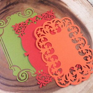 Puede incluir: Tres recortes de papel decorativos en tonos verde, rojo y naranja, dispuestos sobre una superficie de madera. Los recortes presentan intrincados dise&ntilde;os florales y de volutas, con una forma rectangular en verde y formas ovaladas en rojo y naranja.