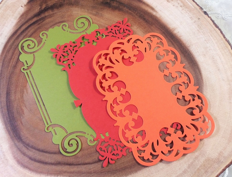 Puede incluir: Tres recortes de papel decorativos en tonos verde, rojo y naranja, dispuestos sobre una superficie de madera. Los recortes presentan intrincados dise&ntilde;os florales y de volutas, con una forma rectangular en verde y formas ovaladas en rojo y naranja.