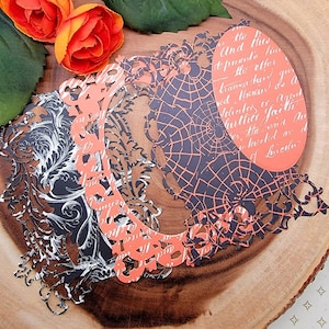May include: A collection of layered paper cutouts in black, orange, and white, featuring ornate frames, spiderwebs, and script. The cutouts are arranged on a wooden surface with orange flowers and green leaves.