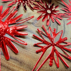 Op de afbeelding: Close-up van verschillende glanzende rode sneeuwvlokornamenten. De ornamenten hebben een metallic afwerking en zijn gerangschikt op een houten oppervlak. De sneeuwvlokontwerpen vari&euml;ren enigszins, met ingewikkelde uitgesneden patronen en puntige randen. Het algemene thema is feestelijk en decoratief.
