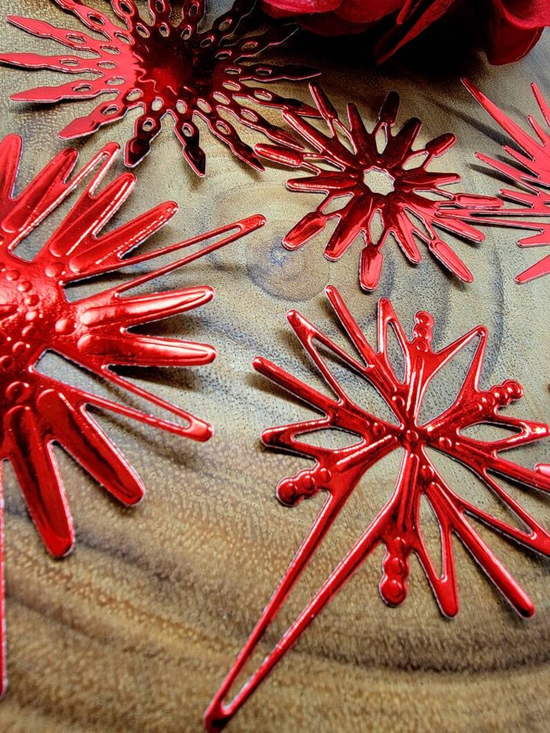 Op de afbeelding: Close-up van verschillende glanzende rode sneeuwvlokornamenten. De ornamenten hebben een metallic afwerking en zijn gerangschikt op een houten oppervlak. De sneeuwvlokontwerpen vari&euml;ren enigszins, met ingewikkelde uitgesneden patronen en puntige randen. Het algemene thema is feestelijk en decoratief.