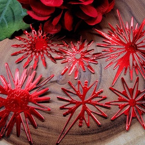 Op de afbeelding: Zes glanzende rode sneeuwvlokornamenten van verschillende ontwerpen zijn gerangschikt op een houten oppervlak. De ornamenten hebben ingewikkelde uitgesneden patronen en een metallic afwerking. Een rode bloem en een groen blad zijn op de achtergrond.