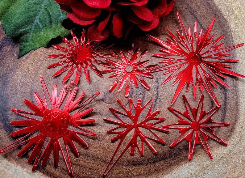 Op de afbeelding: Zes glanzende rode sneeuwvlokornamenten van verschillende ontwerpen zijn gerangschikt op een houten oppervlak. De ornamenten hebben ingewikkelde uitgesneden patronen en een metallic afwerking. Een rode bloem en een groen blad zijn op de achtergrond.