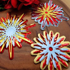 Op de afbeelding: Close-up van drie decoratieve steruitbarstingsornamenten in rood, goud, zilver en wit. De ornamenten hebben een gelaagd ontwerp met een centraal cirkelvormig element en uitstralende punten. De achtergrond is een houten oppervlak met een rode bloem.