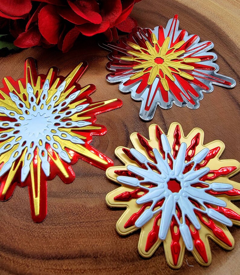 Op de afbeelding: Close-up van drie decoratieve steruitbarstingsornamenten in rood, goud, zilver en wit. De ornamenten hebben een gelaagd ontwerp met een centraal cirkelvormig element en uitstralende punten. De achtergrond is een houten oppervlak met een rode bloem.