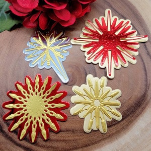 Op de afbeelding: Vier decoratieve steruitbarsting ornamenten in rood, goud en zilver. De ornamenten hebben ingewikkelde ontwerpen en zijn gerangschikt op een houten oppervlak. Een rode bloem staat op de achtergrond.