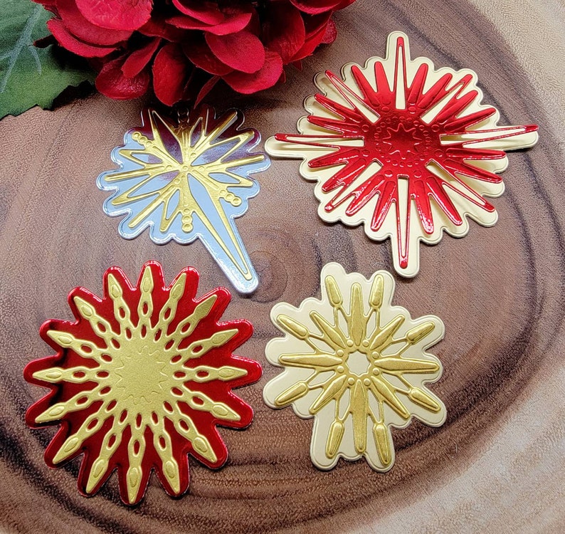 Op de afbeelding: Vier decoratieve steruitbarsting ornamenten in rood, goud en zilver. De ornamenten hebben ingewikkelde ontwerpen en zijn gerangschikt op een houten oppervlak. Een rode bloem staat op de achtergrond.