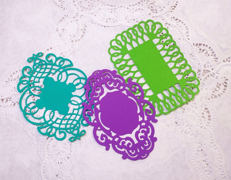 Puede incluir: Tres recortes de papel de colores en verde azulado, morado y verde. Cada recorte tiene un dise&ntilde;o decorativo calado. Los recortes verde azulado y morado tienen patrones ornamentados y arremolinados, mientras que el verde es rectangular con un borde festoneado.