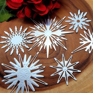 Op de afbeelding: Zes zilveren sneeuwvlok stansvormen in diverse ontwerpen, geplaatst op een houten oppervlak. Een rode bloem en een groen blad zijn op de achtergrond. De sneeuwvlokken hebben ingewikkelde uitgesneden patronen.