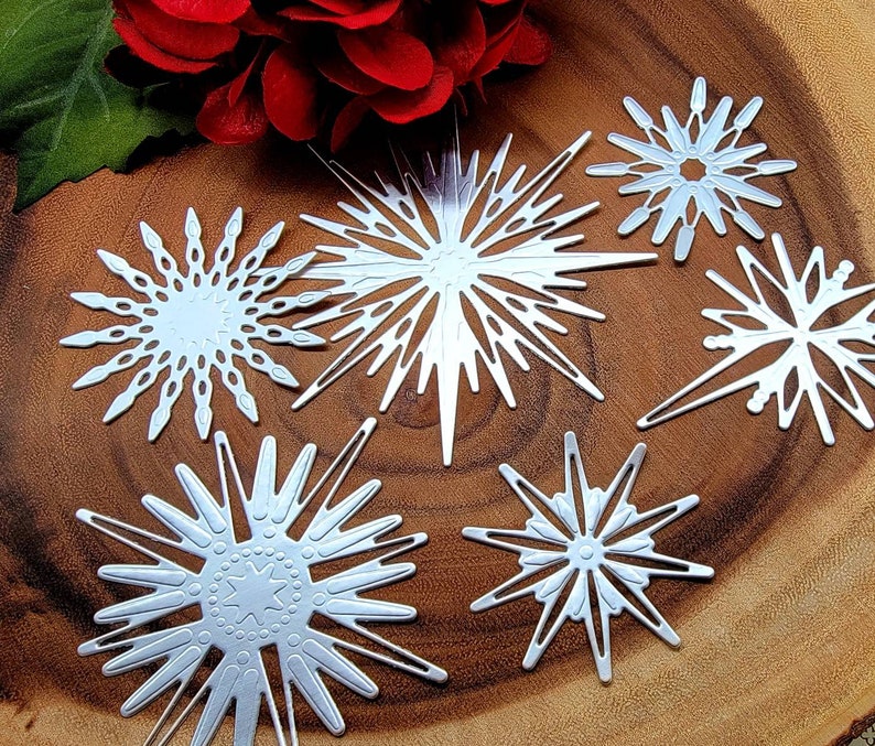 Op de afbeelding: Zes zilveren sneeuwvlok stansvormen in diverse ontwerpen, geplaatst op een houten oppervlak. Een rode bloem en een groen blad zijn op de achtergrond. De sneeuwvlokken hebben ingewikkelde uitgesneden patronen.