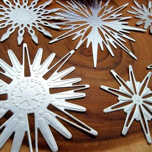 Op de afbeelding: Close-up van verschillende zilveren sneeuwvlokken uitstansingen met ingewikkelde ontwerpen. De sneeuwvlokken vari&euml;ren in grootte en vorm, met patronen van sterren en geometrische vormen. Ze zijn gerangschikt op een warmbruin houten oppervlak.