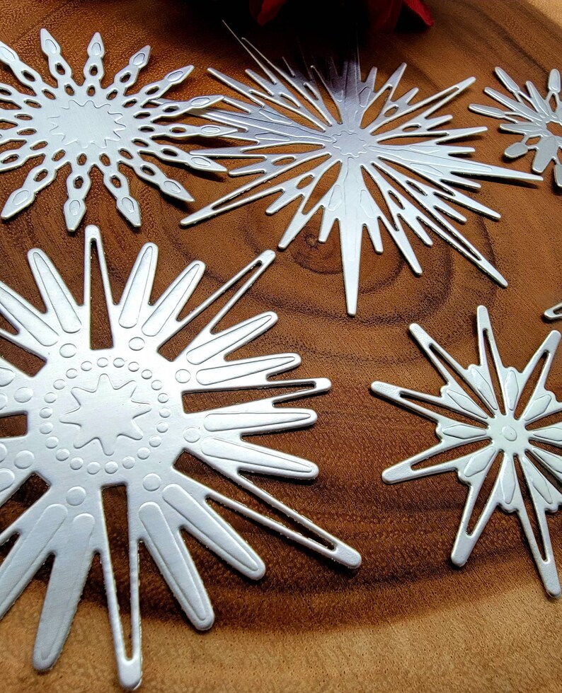 Op de afbeelding: Close-up van verschillende zilveren sneeuwvlokken uitstansingen met ingewikkelde ontwerpen. De sneeuwvlokken vari&euml;ren in grootte en vorm, met patronen van sterren en geometrische vormen. Ze zijn gerangschikt op een warmbruin houten oppervlak.