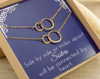 May include: Two gold necklaces with interlocking circles in a blue box. The box has the text "Side by side or miles apart Sisters will be connected by heart."