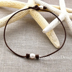May include: A simple brown leather necklace with a single white pearl. The necklace is tied with a knot on each side of the pearl. The necklace is photographed on a light brown fabric background with two white starfish.