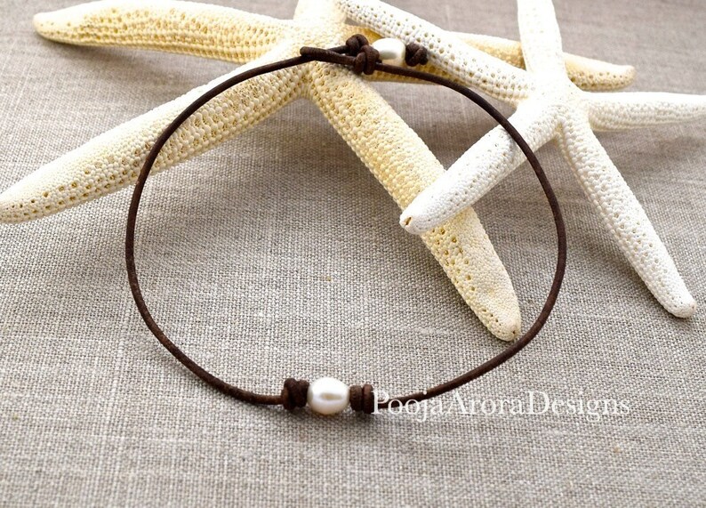 May include: A simple brown leather necklace with a single white pearl. The necklace is tied with a knot on each side of the pearl. The necklace is photographed on a light brown fabric background with two white starfish.
