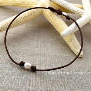 May include: A simple brown leather choker necklace with a single white pearl accent. The necklace is displayed on a textured fabric background.