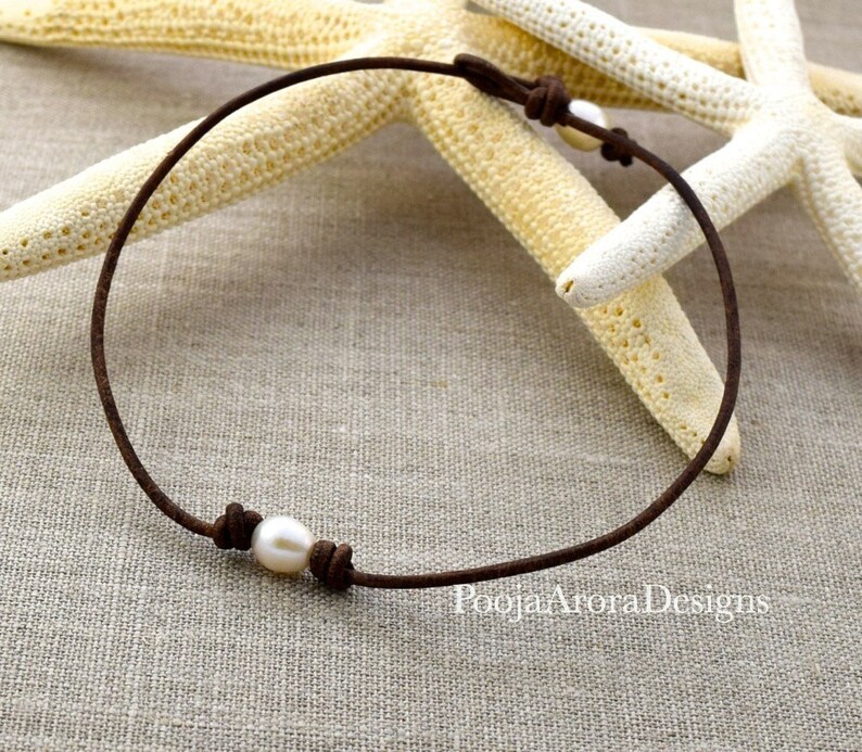 May include: A simple brown leather choker necklace with a single white pearl accent. The necklace is displayed on a textured fabric background.