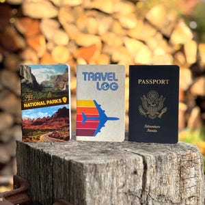 May include: Three travel-themed notebooks. One features a National Parks design, another says "Travel Log" with a plane graphic, and the third is a black passport with "Adventure Awaits" text.