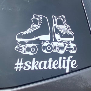 May include: A white vinyl decal featuring a pair of quad roller skates with detailed laces and wheels, positioned above the text #skatelife in a bold, white, sans-serif font. The design is applied to a dark, smooth, reflective surface.