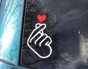 May include: A white vinyl sticker of a hand making the Korean heart sign with a small red heart above it.