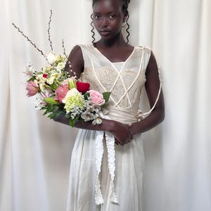 May include: A person wearing a white sleeveless dress with lace detailing and a white sash. They are holding a bouquet of pink, white, and red flowers.
