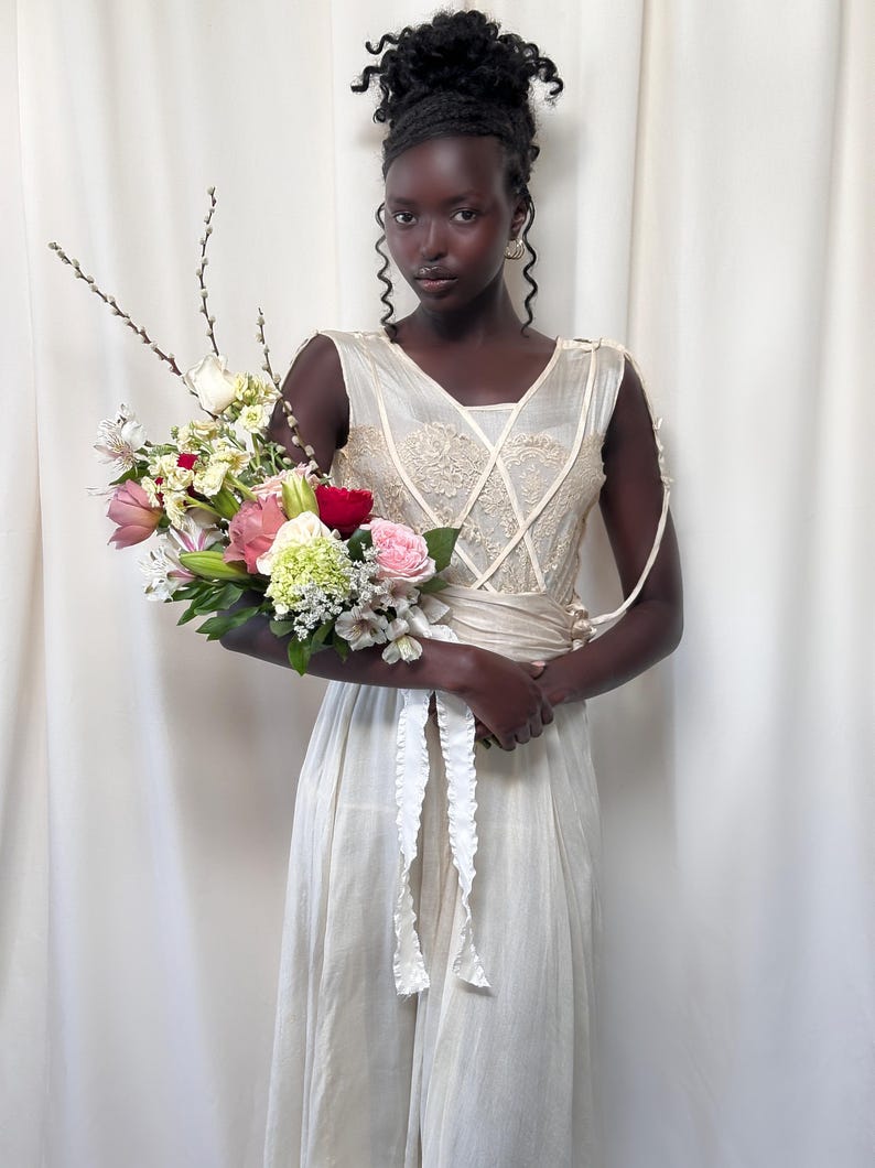 May include: A person wearing a white sleeveless dress with lace detailing and a white sash. They are holding a bouquet of pink, white, and red flowers.