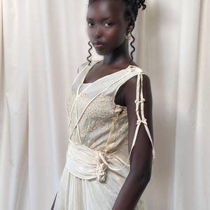 May include: A woman wearing a white sleeveless dress with a lace bodice and a sash tied at the waist. The dress has a vintage style and is made of a sheer fabric.
