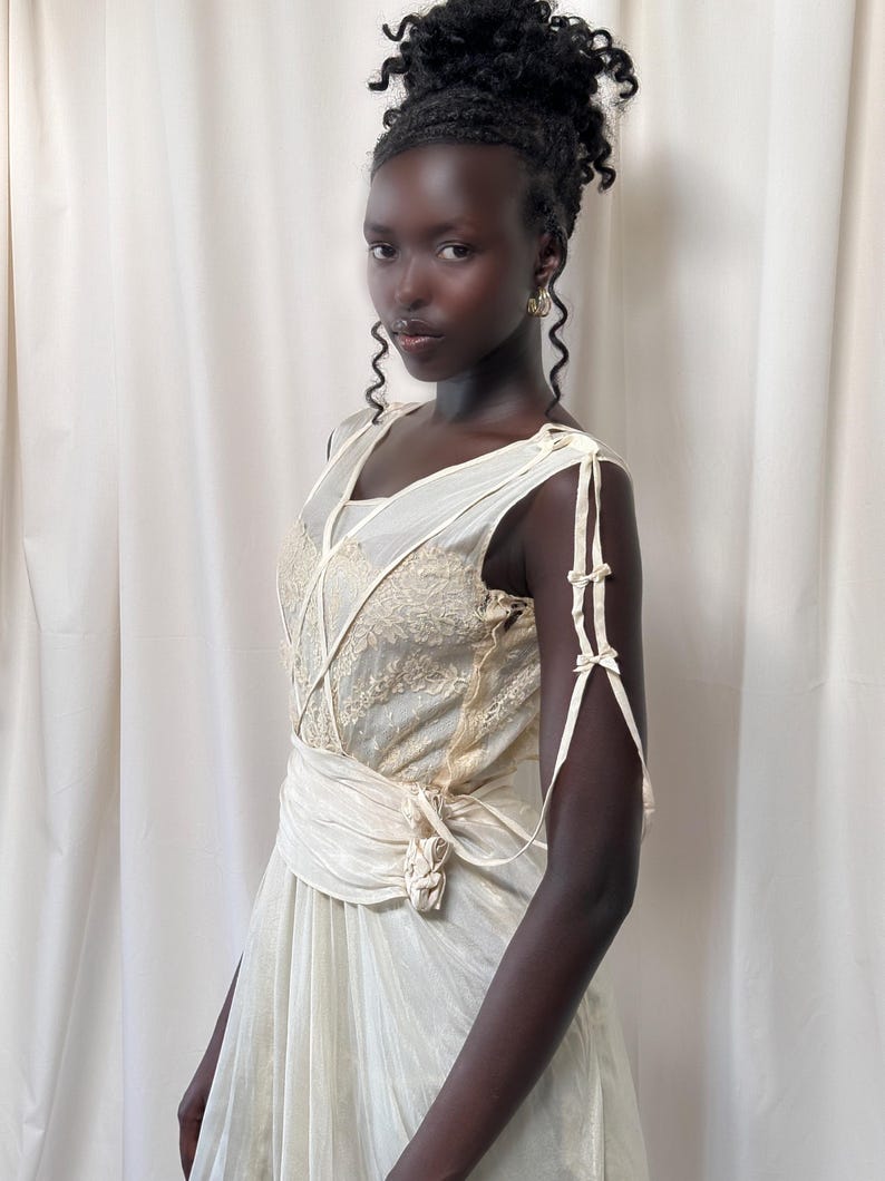 May include: A woman wearing a white sleeveless dress with a lace bodice and a sash tied at the waist. The dress has a vintage style and is made of a sheer fabric.