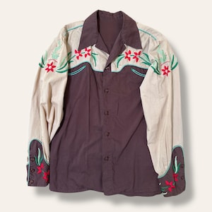 May include: A vintage button-down shirt with a two-tone design. The shirt features a dark brown body and tan sleeves with floral embroidery in red, green, and turquoise. The collar and cuffs are also brown. A classic Western-style shirt.