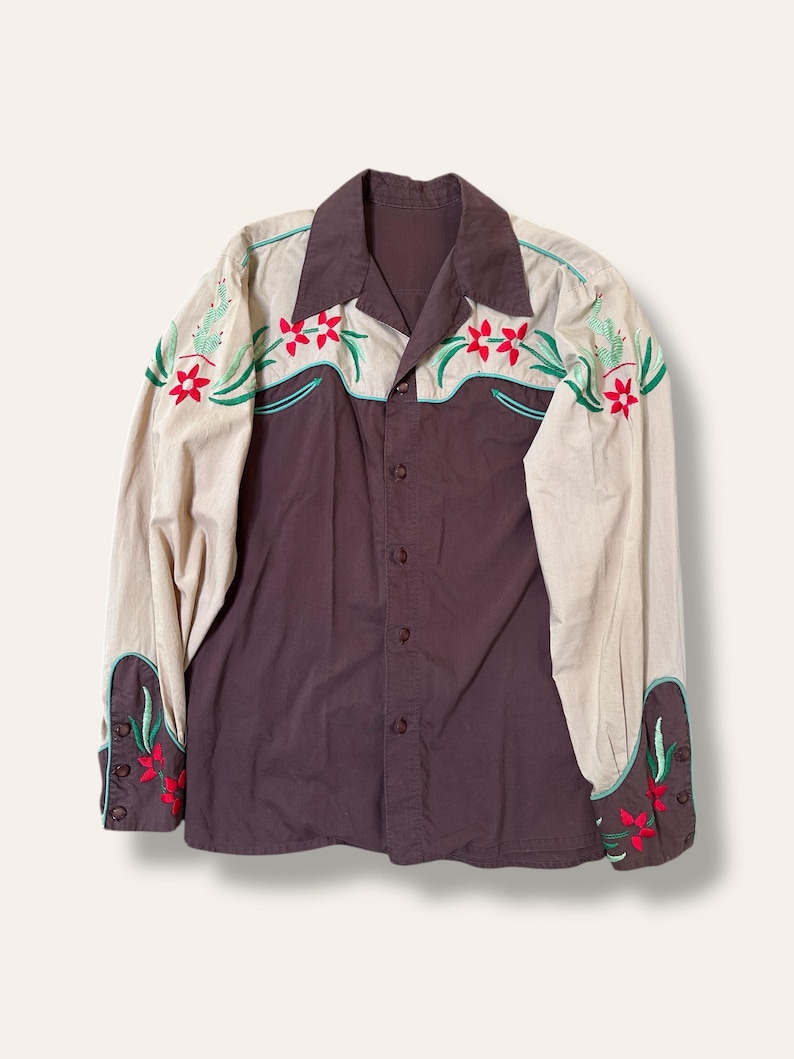 May include: A vintage button-down shirt with a two-tone design. The shirt features a dark brown body and tan sleeves with floral embroidery in red, green, and turquoise. The collar and cuffs are also brown. A classic Western-style shirt.