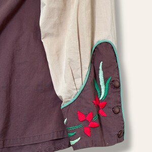 May include: Close-up of a shirt with a color-blocked design. The shirt features a dark brown body and a tan sleeve. The cuff is dark brown with red and green floral embroidery and a teal border. Brown buttons secure the cuff.
