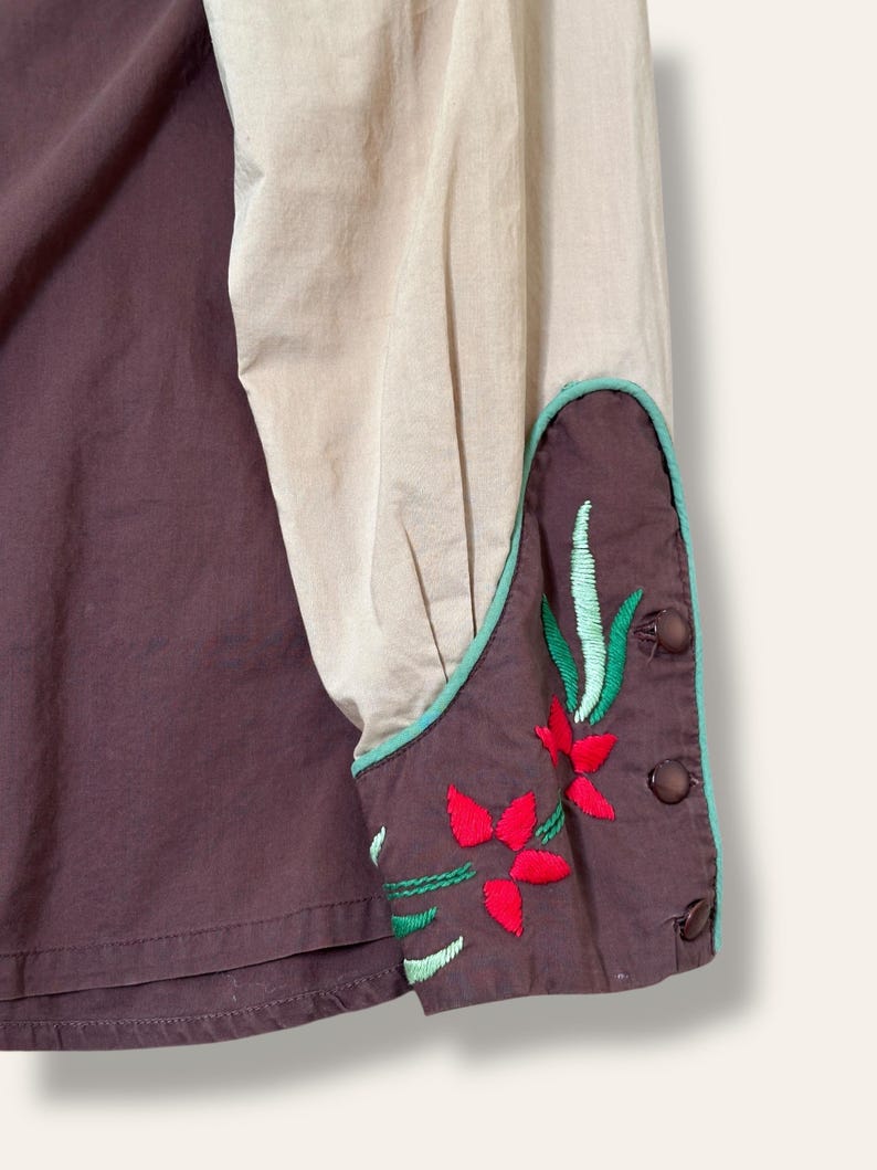 May include: Close-up of a shirt with a color-blocked design. The shirt features a dark brown body and a tan sleeve. The cuff is dark brown with red and green floral embroidery and a teal border. Brown buttons secure the cuff.