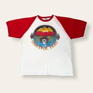 May include: White t-shirt with red sleeves featuring a circular graphic with a skeleton, roses, and a bird. The words "Grateful Dead" are in a circle around the graphic. The shirt has a classic baseball tee style.